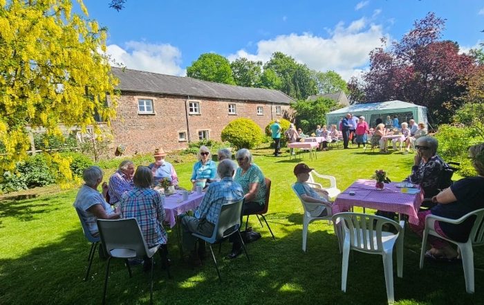 People are sitting around tables in groups on a green lawn in a garden in the sunshine