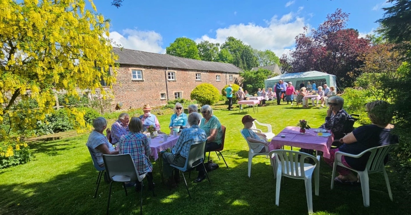 People are sitting around tables in groups on a green lawn in a garden in the sunshine