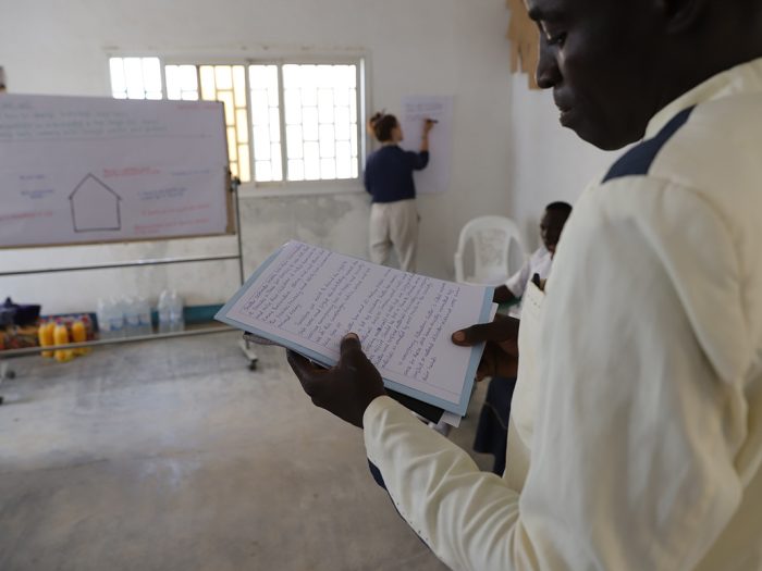 Man in white reading from a piece of paper in a classroom