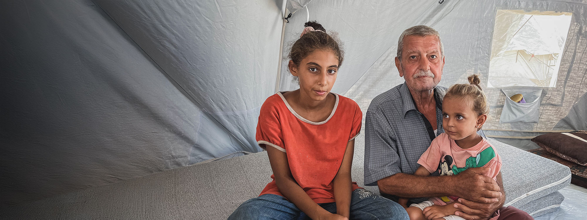 man sits with two daughters on a mattress inside an emergency tent