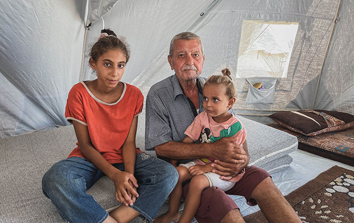 man sits with two daughters on a mattress inside an emergency tent