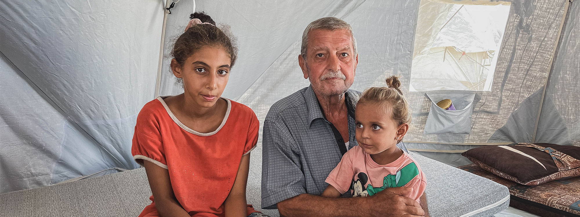 man sits with two daughters on a mattress inside an emergency tent