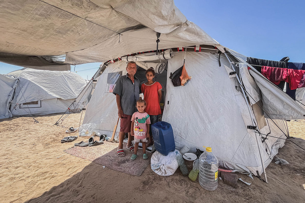 man stands with two daughters outside emergency tent wide angle