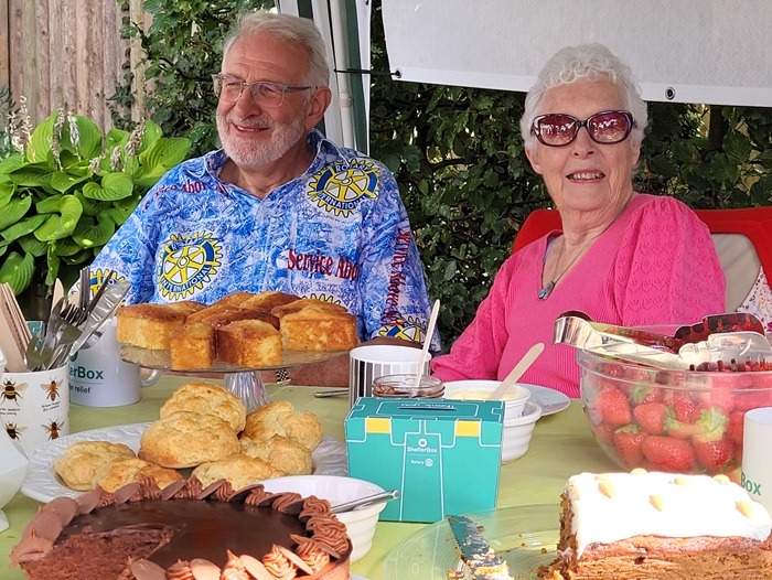 A man and a woman are sitting at a table covered with cakes, in the middle is a Tea in a Tent collection box